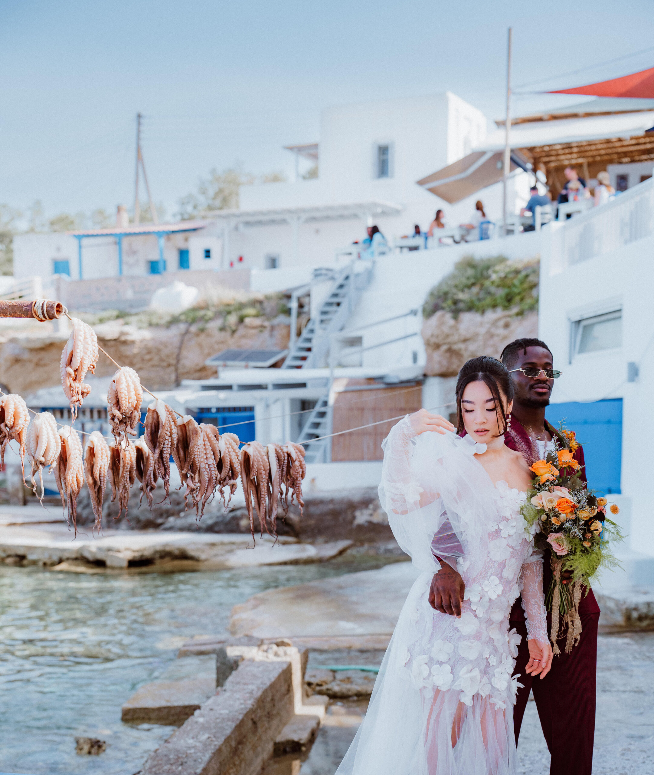 Bride and Groom pose at seaside Greek Village with octopus drying on the line, showcasing a multicultural wedding with modern floral details and costal charm