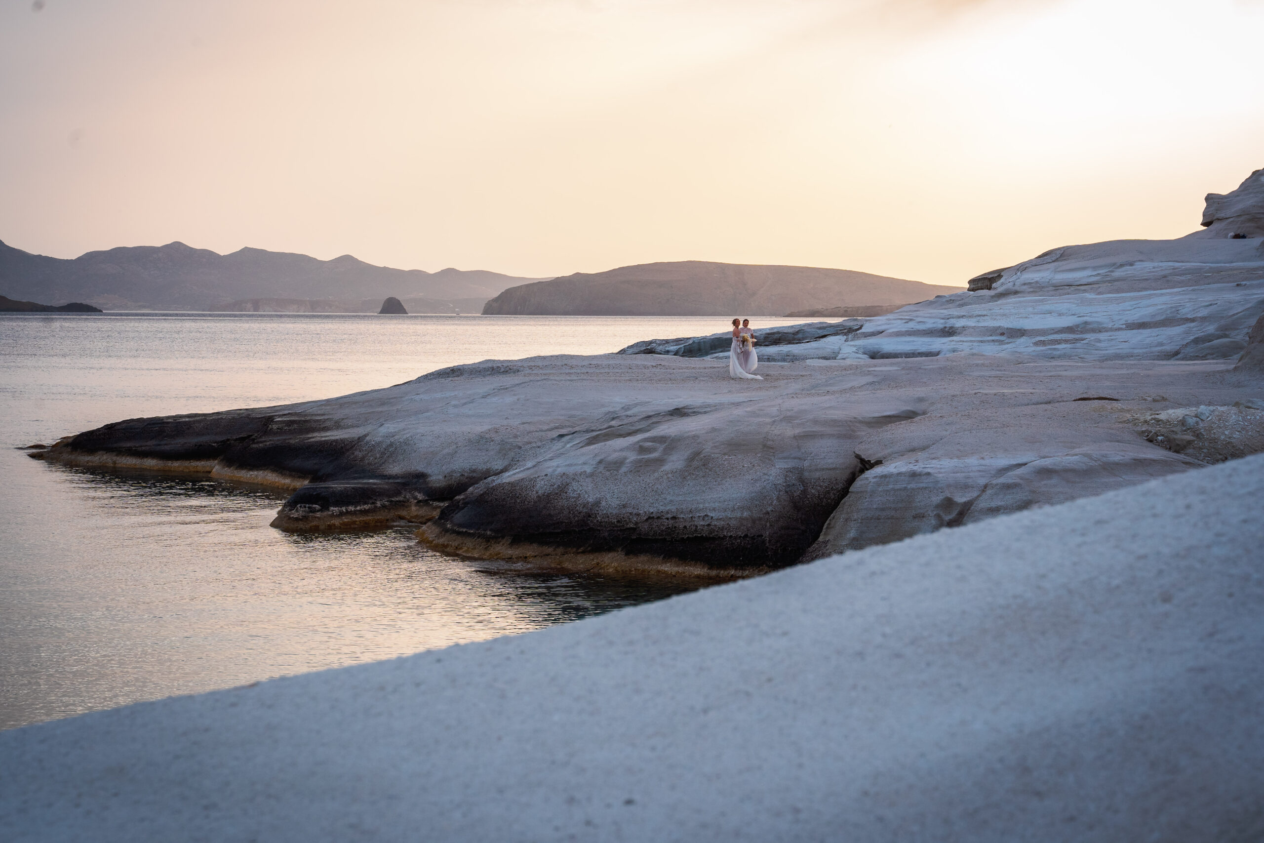 Two brides standing on the white cliffs of Sarakiniko Beach, Milos Greece during a romantic same-sex elopement editorial captured by Chi Ramos Studio.